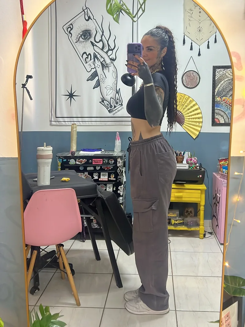 Milena Fuentes em pé no estúdio, fotografada no espelho em arco, com cadeira rosa e desenhos ao fundo