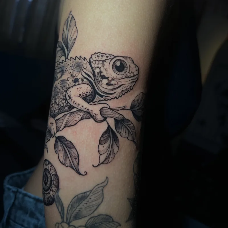 Tatuagem em tinta preta de camaleão sobre um galho, com folhas, contornos e sombreamento em pontos