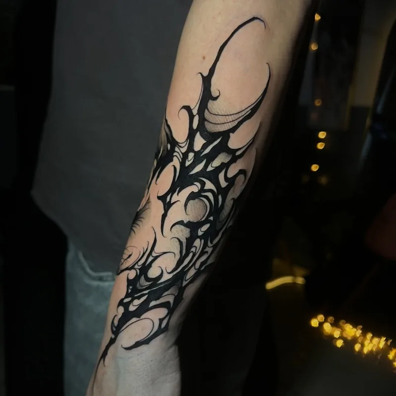 Tatuagem abstrata em tinta preta no antebraço, com formas curvas, pontas alongadas e espaços de pele entre os traços