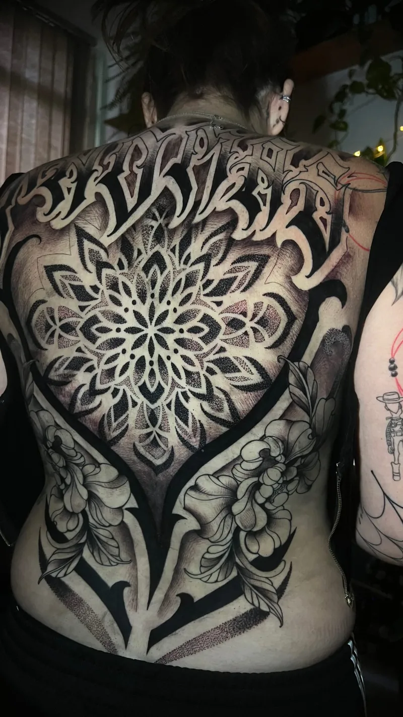 Tatuagem em preto cobrindo as costas, com mandala central, flores, folhas e áreas preenchidas em pontilhismo