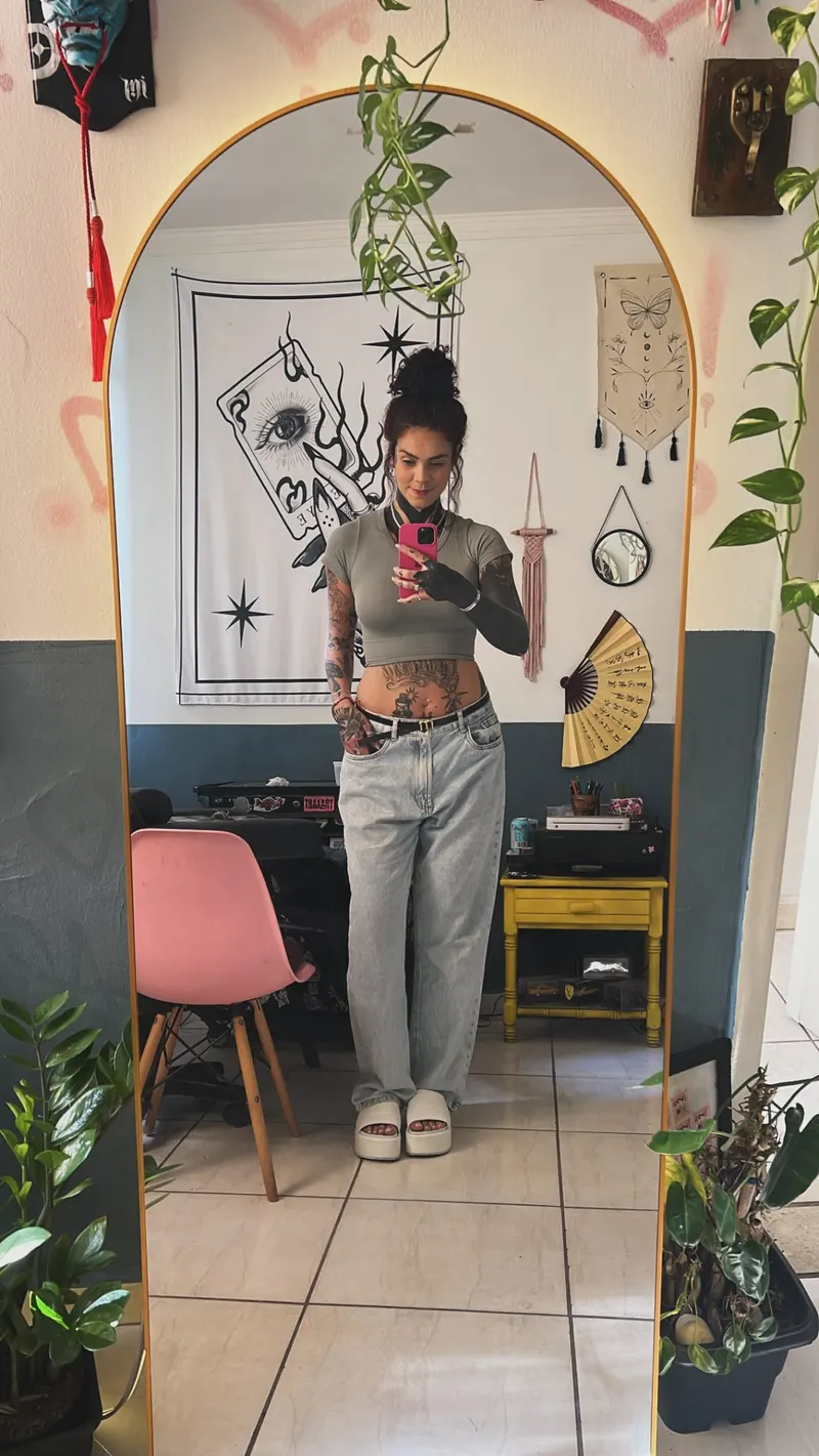 Estúdio de tatuagem visto pelo espelho em arco, com cadeira rosa, plantas e quadros nas paredes